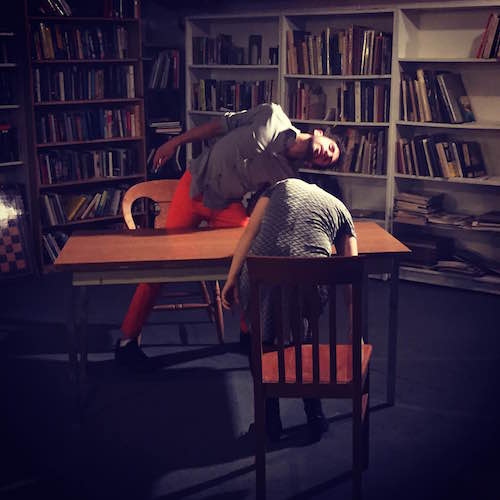 Two dancers shake their head around a library table. 